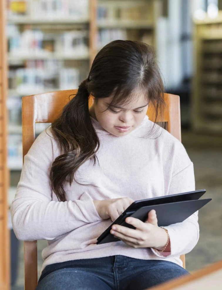 Une jeune personne assise dans une bibliothèque et regardant une tablette